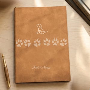 May include: A tan notebook with a smooth, matte texture featuring a white line-art dog icon at the top, a horizontal row of six light-colored paw prints across the center, and the text "Pet's Name" printed in a white script font at the bottom.