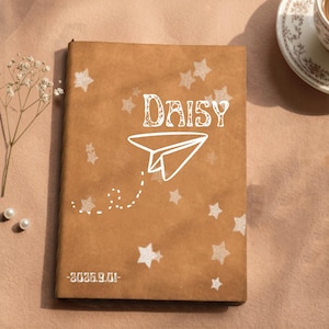 Może przedstawiać: Brązowy notatnik z białą grafiką papierowego samolotu, napisem "DAISY" stylizowaną czcionką oraz rozrzuconymi białymi gwiazdkami. Data "2026.9.01" jest nadrukowana na biało w lewym dolnym rogu prostokątnej okładki tego przedmiotu.