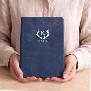 Op de afbeelding: Een marineblauw notitieboek met een getextureerde kaft, voorzien van een witte afbeelding van hertengeweien rondom de letter K en de naam KATIE in witte hoofdletters eronder. Het notitieboek heeft een rechthoekige vorm met afgeronde hoeken.