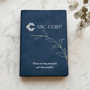 May include: A dark blue rectangular notebook featuring a white geometric logo, the text ABC CORP., a thin gold line drawing of a leafy branch, and the printed phrase Focus on the powerful, not the painful. The cover has a smooth, matte finish.