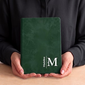 Könnte beinhalten: Ein dunkelgrünes Notizbuch mit texturiertem Einband, das den weiß gedruckten Namen Madeline vertikal neben einem großen weißen Buchstaben M zeigt, welcher ein kleines Blattmotiv in seine Struktur auf der Vorderseite integriert.