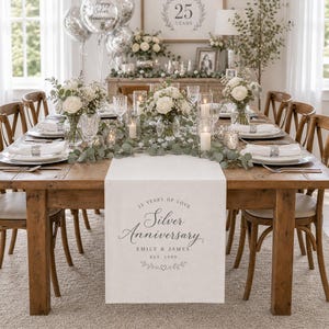 May include: A wooden dining table set for a 25th anniversary with a white runner featuring the text 25 YEARS OF LOVE Silver Anniversary EMILY & JAMES EST. 1999. The table includes white floral centerpieces, candles, and place settings.