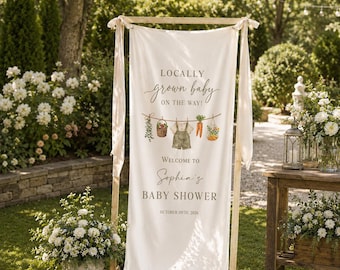 May include: A vertical cream-colored fabric banner suspended from a wooden frame. The text reads LOCALLY grown baby ON THE WAY! WELCOME TO Sophia's BABY SHOWER OCTOBER 09TH, 2026. A graphic shows a clothesline with a basket, baby overalls, and carrots.
