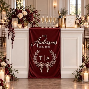 Op de afbeelding: Een dieprode stoffen tafelloper met witte opdruk The Andersons EST. 2026, gecentreerd boven een decoratief lauwerkransontwerp met een strik en de initialen T A. De loper ligt over een witte tafel met bloemendecoratie.