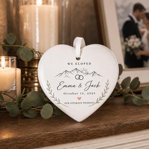 May include: A white heart-shaped ceramic ornament featuring a black line drawing of mountains, a sun, and two interlocking rings. The text reads WE ELOPED, Emma & Jack, October 12, 2025, and OUR INTIMATE WEDDING, with a small pink heart accent.