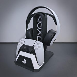 May include: A black and white gaming accessory stand featuring a vertical pillar with the PlayStation symbols triangle, circle, cross, and square. The stand holds a white and black wireless controller and a pair of white and black over-ear headphones.