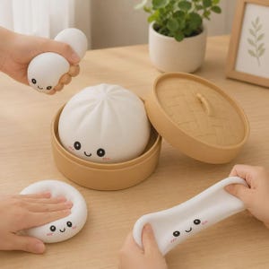 May include: A set of white, soft, squishy dumpling-shaped toys with smiling faces, including one inside a tan, round, lidded bamboo-style steamer basket. The toys are flexible and can be squeezed, flattened, or stretched by hand while maintaining their shape.
