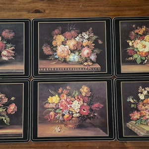 May include: A set of six rectangular table mats featuring floral still life paintings. Each mat has a black border and displays a unique arrangement of colourful flowers in baskets or vases, set against a dark background on a wooden surface.