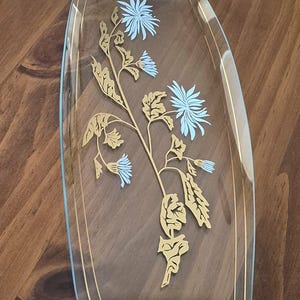 May include: A rectangular glass serving tray with curved sides, featuring a gold and light blue floral design. The tray has a wooden handle or accent piece at both the top and bottom edges, and thin gold lines running along the length of the glass.