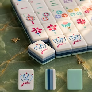 May include: A close-up of a Mahjong set with white tiles featuring colorful floral and geometric designs. Several tiles display the word "joker" in a script font. The tiles are arranged on a green marble surface.
