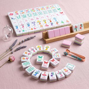 May include: A complete Mahjong set with white tiles featuring colorful floral and bird designs. The set includes pink tiles, a wooden rack, and decorative tongs. The tiles are arranged on a pink surface, suggesting a game-themed product.