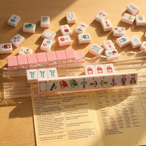 May include: A Mahjong set with pink and white tiles, featuring various symbols and illustrations. The tiles are arranged on a wooden surface, with a clear plastic rack and a rule sheet visible.