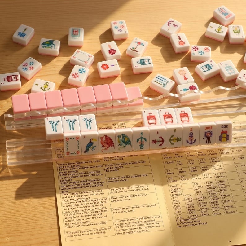 May include: A Mahjong set with pink and white tiles, featuring various symbols and illustrations. The tiles are arranged on a wooden surface, with a clear plastic rack and a rule sheet visible.