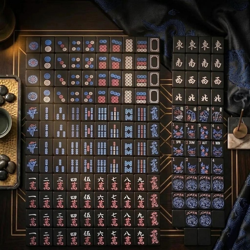 May include: A set of black Mahjong tiles featuring engraved characters, numbers, and symbols in red, blue, and white. The tiles include suits of dots, bamboo, and characters, along with wind tiles, dragon tiles, flower tiles, and Joker tiles.
