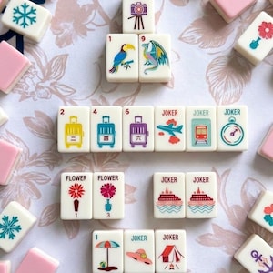 May include: A collection of white and pink Mahjong tiles with various colorful illustrations. The tiles feature images of flowers, suitcases, a toucan, a dragon, and other symbols. Some tiles have the word "JOKER" printed on them.