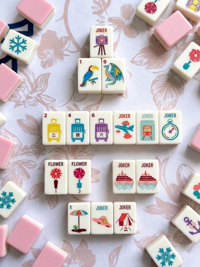 May include: A collection of white and pink Mahjong tiles with various colorful illustrations. The tiles feature images of flowers, suitcases, a toucan, a dragon, and other symbols. Some tiles have the word "JOKER" printed on them.
