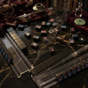 May include: A Mahjong set featuring black tiles with red, blue, and white characters and symbols. The tiles are arranged on a black marble surface with gold veining, accompanied by clear acrylic tile racks, a calligraphy brush, and a small glass bottle.