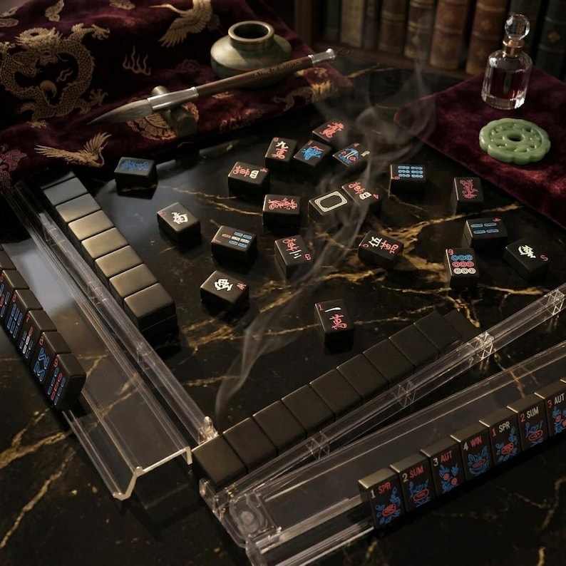 May include: A Mahjong set featuring black tiles with red, blue, and white characters and symbols. The tiles are arranged on a black marble surface with gold veining, accompanied by clear acrylic tile racks, a calligraphy brush, and a small glass bottle.