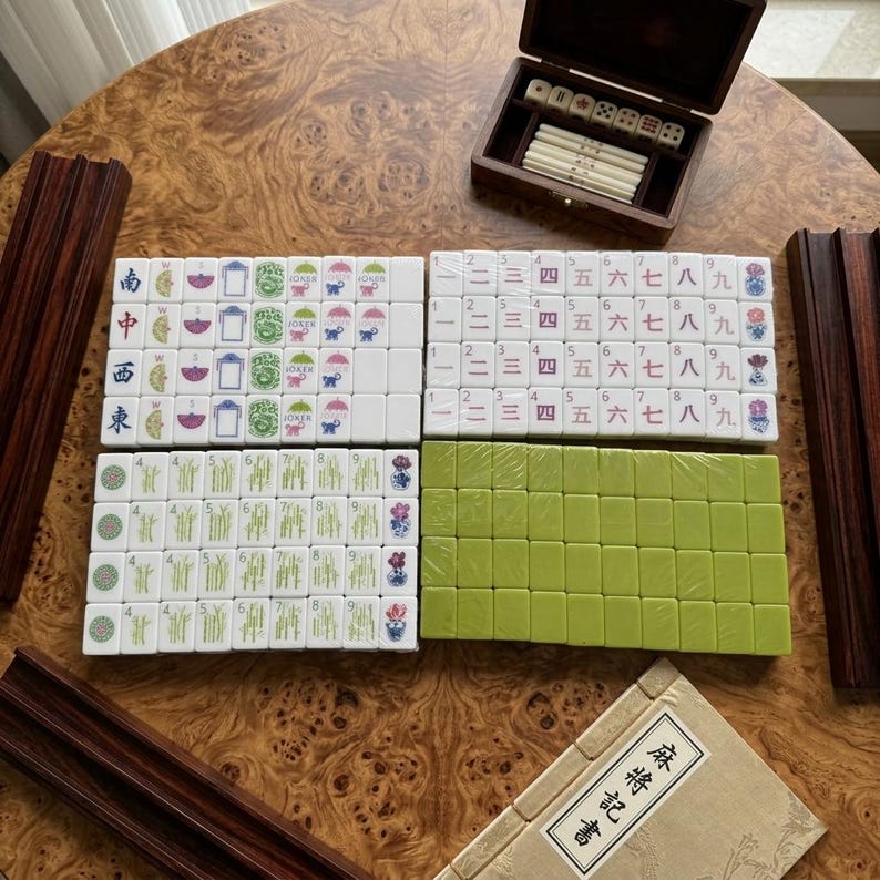 May include: A Mahjong set featuring white tiles with green, blue, and red character, bamboo, and flower designs, alongside a set of solid green tiles. The set includes a wooden storage box with dice and counting sticks, and a manual with Japanese text.