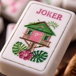 May include: A white rectangular Mahjong tile featuring the word JOKER in pink text at the top. The centre displays a green and pink treehouse illustration with the letters PS on the door, surrounded by green palm leaves and a pink hibiscus flower.