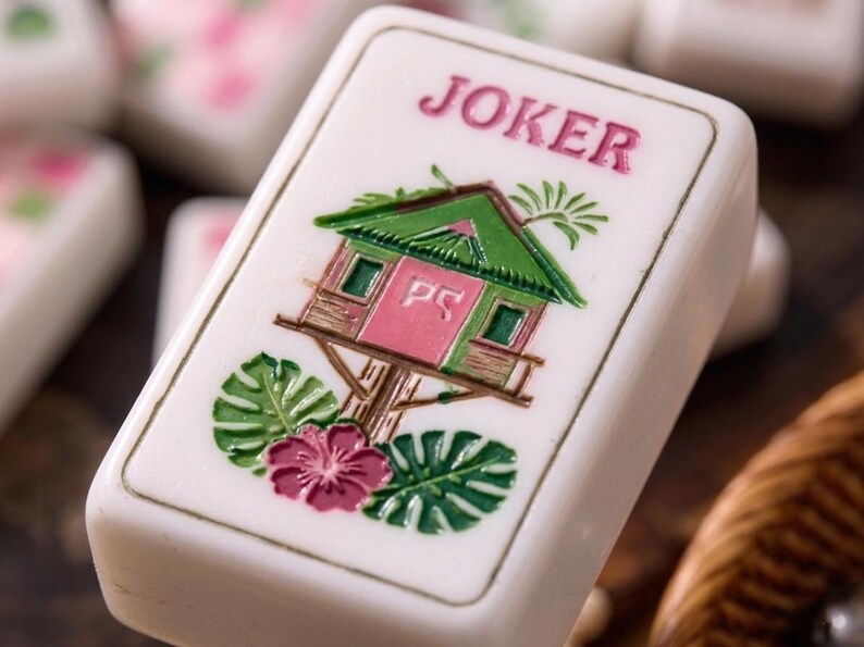 May include: A white rectangular Mahjong tile featuring the word JOKER in pink text at the top. The centre displays a green and pink treehouse illustration with the letters PS on the door, surrounded by green palm leaves and a pink hibiscus flower.