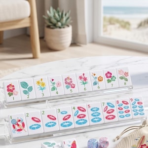May include: A set of white Mahjong tiles with floral and water-themed illustrations, including hibiscus, leaves, and life preservers. The tiles are arranged in clear acrylic racks, with dice and a small bag visible.