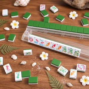 K&ouml;nnte beinhalten: Ein komplettes Mahjong-Set mit gr&uuml;nen und wei&szlig;en Spielsteinen, mit tropischen Illustrationen und Zahlen. Das Set enth&auml;lt ein durchsichtiges Kunststoff-Spielsteinregal, Muscheln und wei&szlig;e Blumen, alles auf einer Holzoberfl&auml;che angeordnet.