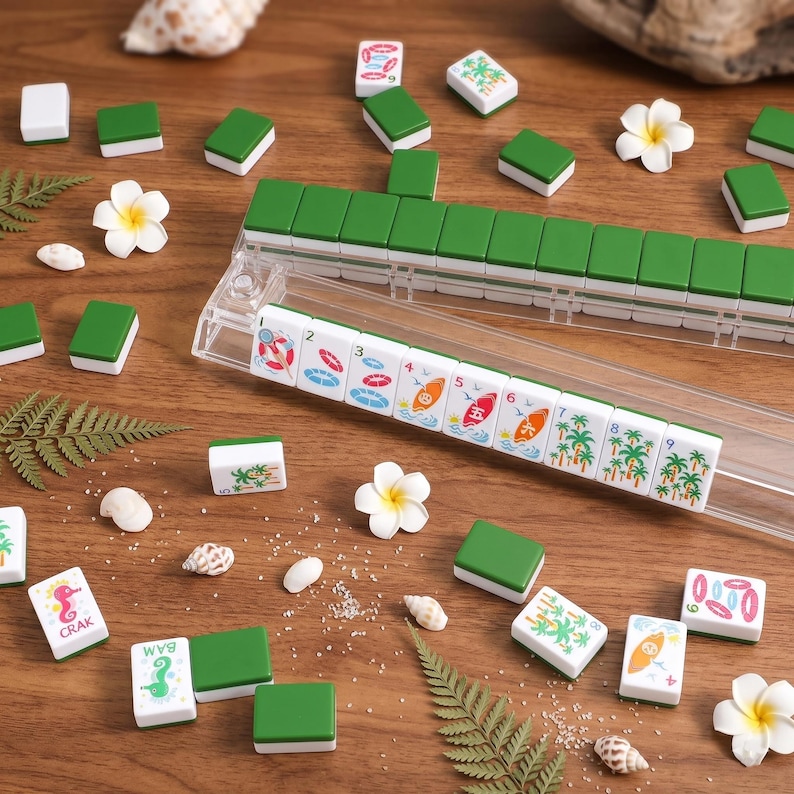 K&ouml;nnte beinhalten: Ein komplettes Mahjong-Set mit gr&uuml;nen und wei&szlig;en Spielsteinen, mit tropischen Illustrationen und Zahlen. Das Set enth&auml;lt ein durchsichtiges Kunststoff-Spielsteinregal, Muscheln und wei&szlig;e Blumen, alles auf einer Holzoberfl&auml;che angeordnet.