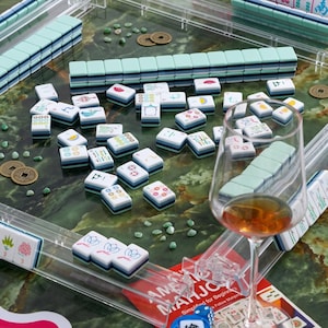 May include: A complete Mahjong set with white and teal tiles, arranged on a green marble-patterned surface. Includes a glass of amber liquid, dice, and decorative coins. The set is designed for beginners.