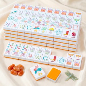 May include: A complete Mahjong set with white tiles featuring colorful floral and symbol designs, stacked neatly. The tiles have orange edges. Additional game pieces include orange stones and a green dragonfly.