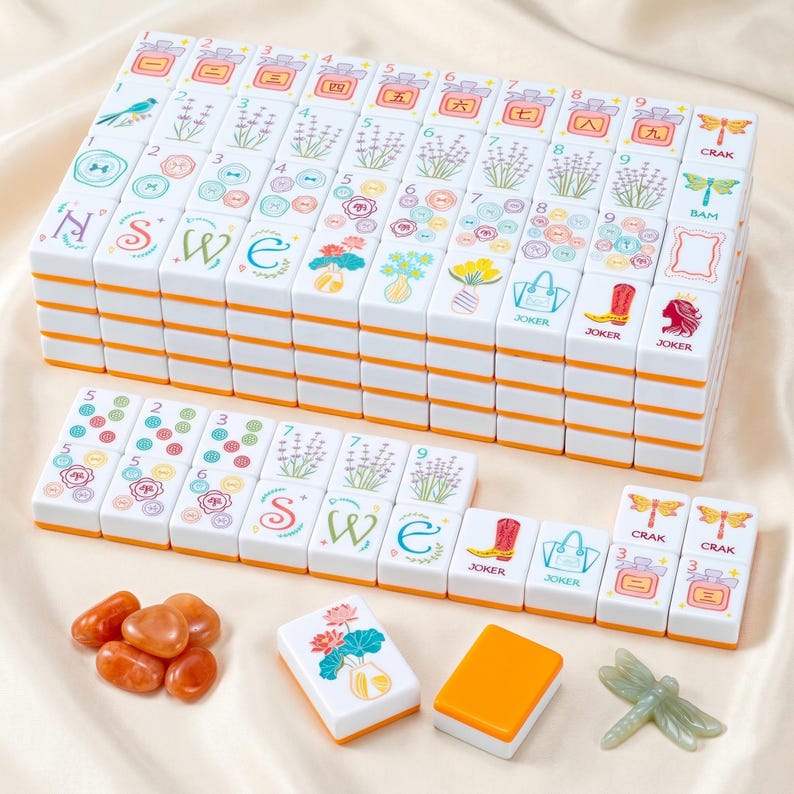 May include: A complete Mahjong set with white tiles featuring colorful floral and symbol designs, stacked neatly. The tiles have orange edges. Additional game pieces include orange stones and a green dragonfly.
