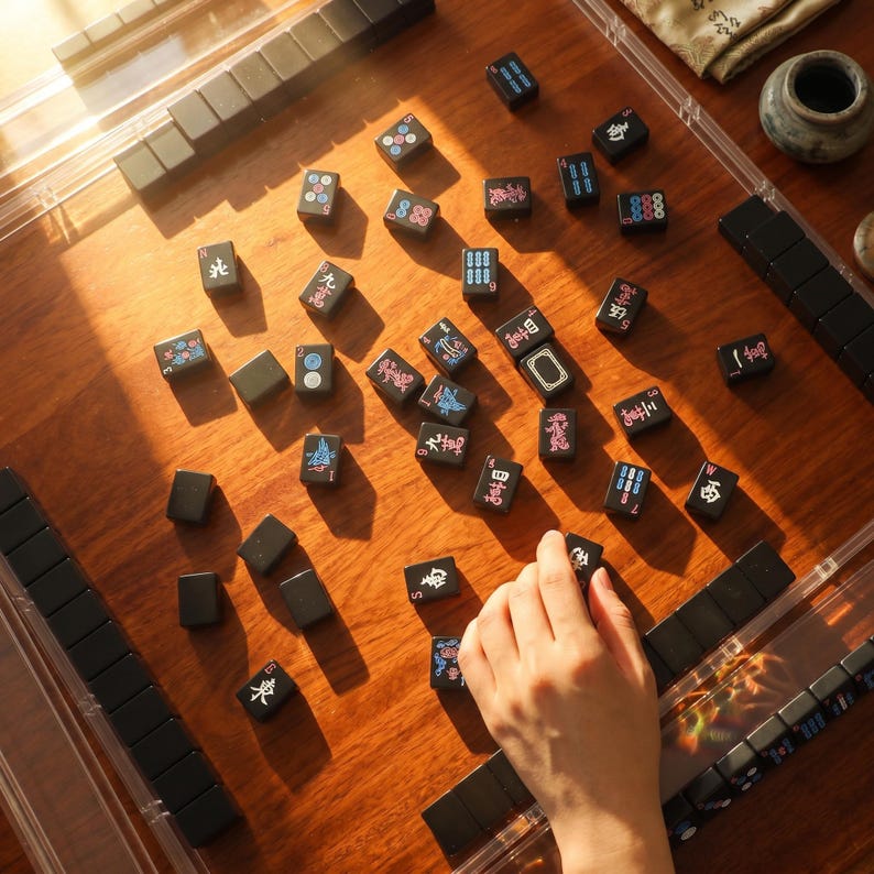 May include: A set of black Mahjong tiles with colorful engraved characters and symbols scattered across a wooden surface, with some tiles arranged in clear plastic racks and one tile being held by a hand during gameplay.
