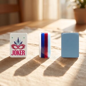 May include: Three rectangular glass game pieces. One is white with a red and teal mask design and the word "JOKER". Another is striped with red, white, and blue. The third is solid light blue. They are on a textured surface.