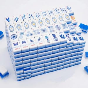 May include: A large set of white mahjong tiles stacked in a rectangular block. Each tile features blue and white illustrations of vases, flowers, butterflies, birds, and fish, along with red text labels for directions and numbers in blue ink.