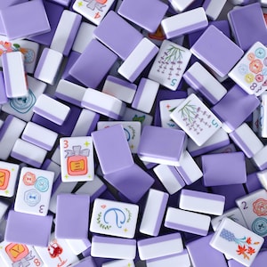 May include: Overhead shot of a collection of purple and white Mahjong tiles. Some tiles have illustrations of flowers, numbers, and symbols. The tiles are scattered, creating a visually appealing pattern.