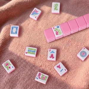 May include: Pink and white Mahjong tiles scattered on a pink surface. The tiles feature various symbols and characters in green, blue, and red. A set of pink tiles is also visible, suggesting a complete game set.
