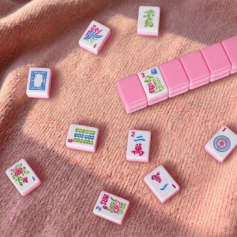 May include: Pink and white Mahjong tiles scattered on a pink surface. The tiles feature various symbols and characters in green, blue, and red. A set of pink tiles is also visible, suggesting a complete game set.
