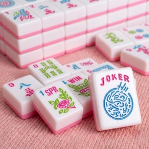 May include: White and pink Mahjong tiles with various symbols and characters. The tiles have pink edges and are stacked in rows. Some tiles are scattered, displaying the word "JOKER" and other symbols in green, pink, and blue.