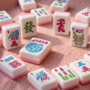 May include: A collection of white and pink Mahjong tiles with various symbols and characters. Some tiles feature numbers, while others display dragons, flowers, and the word "JOKER". The tiles are arranged on a pink surface.