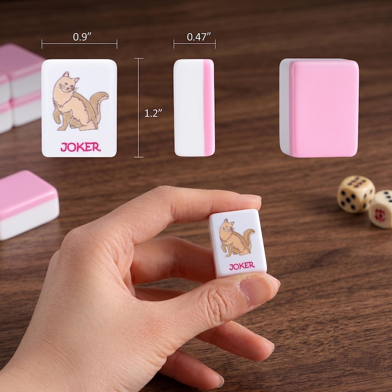 May include: White and pink Mahjong tiles featuring a cat illustration and the word "JOKER". The tiles measure approximately 2.3 cm x 3 cm x 1.2 cm. Two dice are visible. The tiles are on a wooden surface.
