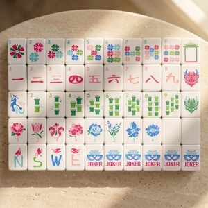 May include: A complete set of white Mahjong tiles with various colorful floral and Chinese symbol designs. The tiles feature numbers, bamboo, and joker tiles. The set is arranged on a light-colored surface.