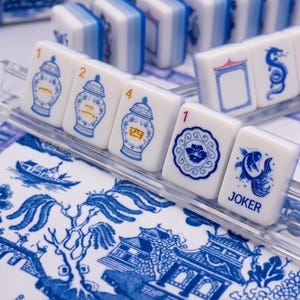 Puede incluir: Un juego de fichas de Mahjong blancas y azules con varias ilustraciones, incluyendo jarrones y peces, dispuestas sobre una bandeja transparente. Las fichas se colocan sobre una tela estampada en azul y blanco, creando una est&eacute;tica cl&aacute;sica.