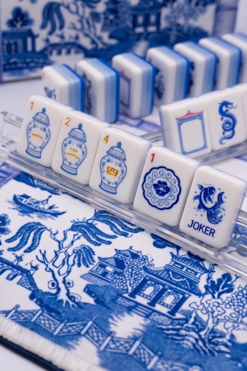 Puede incluir: Un juego de fichas de Mahjong blancas y azules con varias ilustraciones, incluyendo jarrones y peces, dispuestas sobre una bandeja transparente. Las fichas se colocan sobre una tela estampada en azul y blanco, creando una est&eacute;tica cl&aacute;sica.