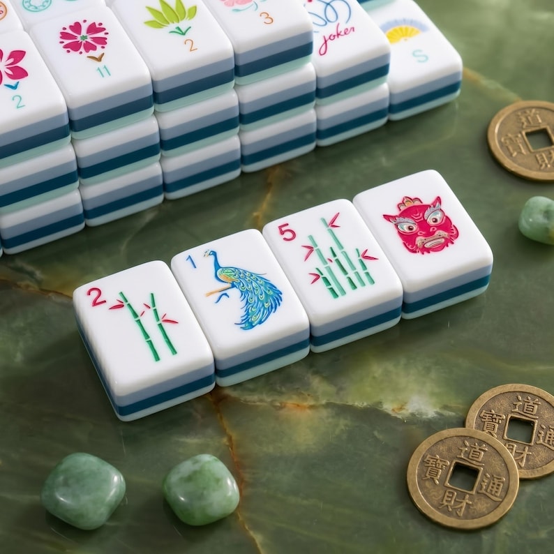 K&ouml;nnte beinhalten: Ein Satz wei&szlig;-blauer Mahjong-Steine mit verschiedenen Symbolen und Zahlen, darunter ein Pfau, Bambus und ein rotes Gesicht. Zwei chinesische M&uuml;nzen und gr&uuml;ne Steine liegen auf einer gr&uuml;nen Oberfl&auml;che.