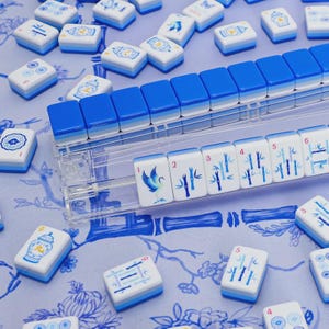 K&ouml;nnte beinhalten: Eine Sammlung wei&szlig;-blauer Mahjong-Steine mit verschiedenen Illustrationen und Zahlen. Die Steine sind auf einer blau gebl&uuml;mten Oberfl&auml;che angeordnet, einige in einem transparenten Kunststoffhalter. Die Motive zeigen Bambus, V&ouml;gel und andere traditionelle Designs.