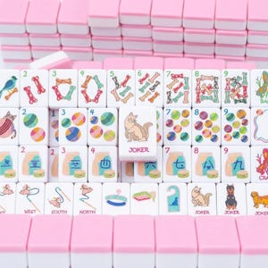 May include: A pink and white Mahjong tile set. The tiles feature various colorful illustrations, including bones, birds, and cats. One tile is labeled "JOKER". The set is arranged in rows, with pink-topped tiles stacked in the background.