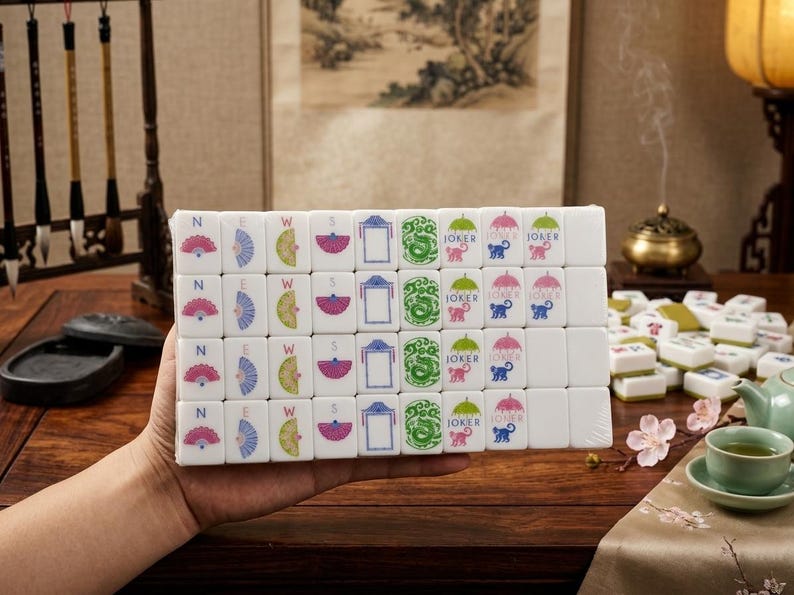 May include: A set of white rectangular Mahjong tiles arranged in a grid, featuring colorful engraved designs including fans, the letters N, E, W, S, blue frames, green dragons, and pink or blue monkeys labeled with the word JOKER and umbrella icons.