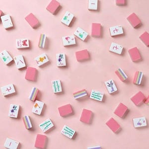 May include: A collection of pink and white Mahjong tiles scattered on a pink surface. The tiles feature various designs, including butterflies, flowers, and numbers. Some tiles are solid pink, while others have colorful stripes.