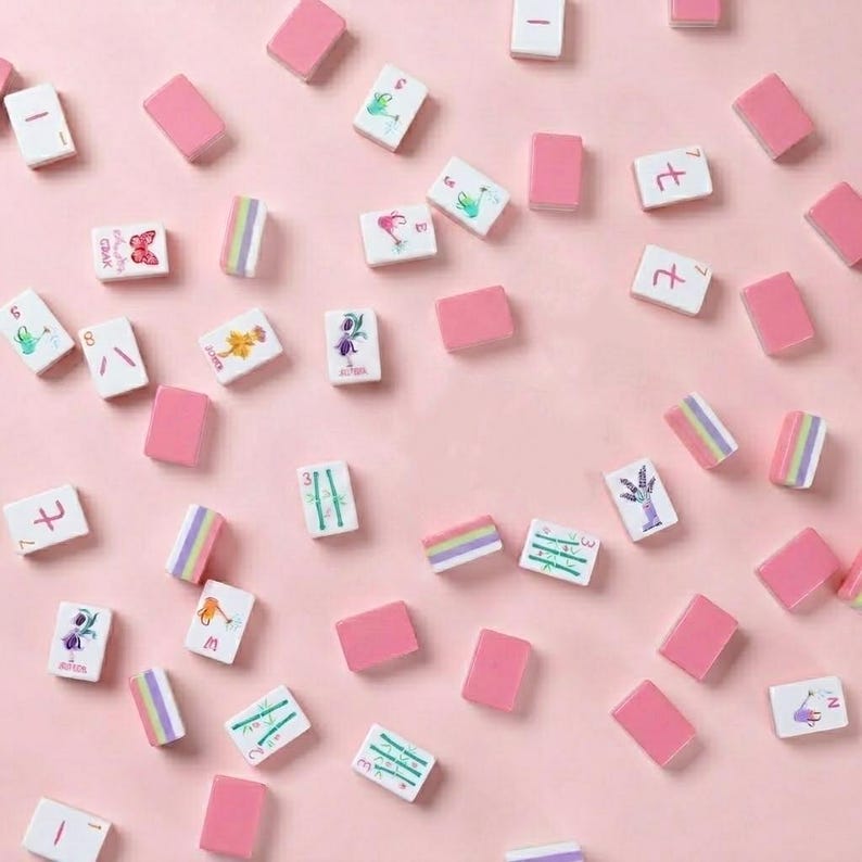 May include: A collection of pink and white Mahjong tiles scattered on a pink surface. The tiles feature various designs, including butterflies, flowers, and numbers. Some tiles are solid pink, while others have colorful stripes.