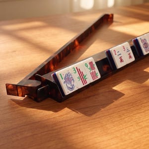 May include: A brown and amber-colored Mahjong tile holder with several white tiles featuring red, green, and blue designs. The tiles are arranged in a row, with the holder's angled arm extending upwards. The set is on a wooden surface.